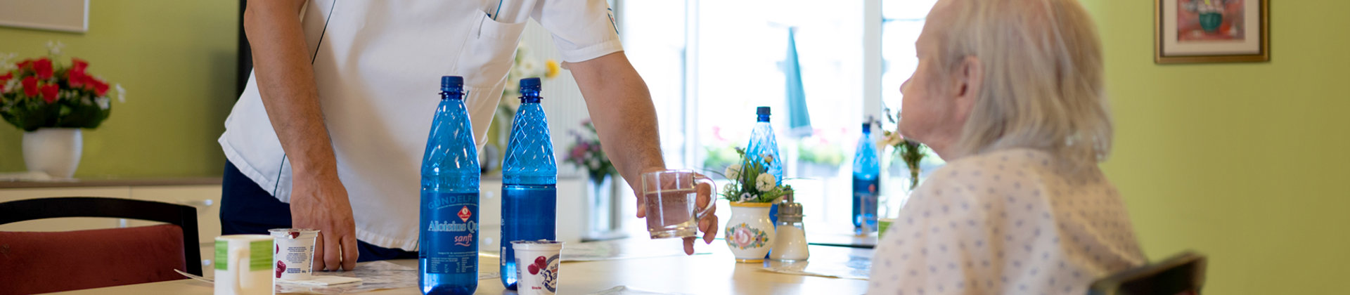 Bild | Pfleger reicht Seniorin ein Glas Wasser am Esstisch